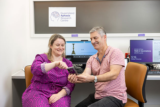 A man and a woman smiling and interacting with a tablet in a medical office with posters on the wall, including one for the Queensland Aphasia Research Centre and another displaying information about COVID-19 vaccinations.