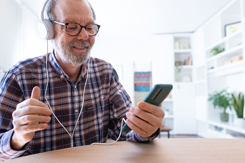 Senior man listening to music and dancing using mobile phone and headphones at home living room