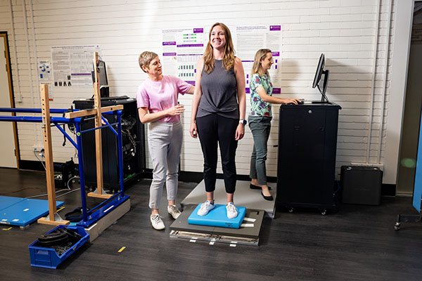 A patient stands on a mat that measures pressure while two researchers record the findings